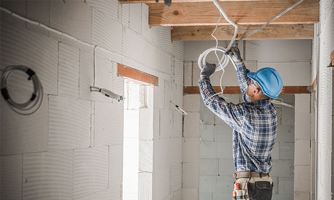 Electrician in a blue hard hat installs wiring in a hallway under construction, surrounded by unfinished walls and wooden beams.