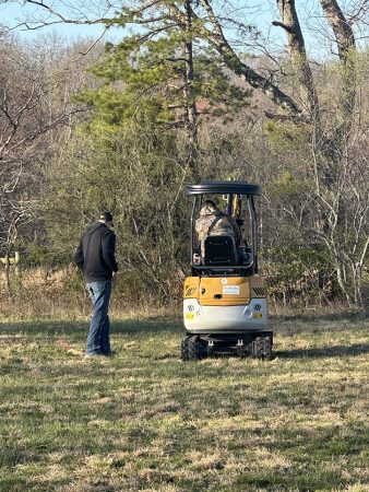 A man in a black jacket stands near a yellow loader, while another person in camouflage operates it in a grassy area surrounded by trees.