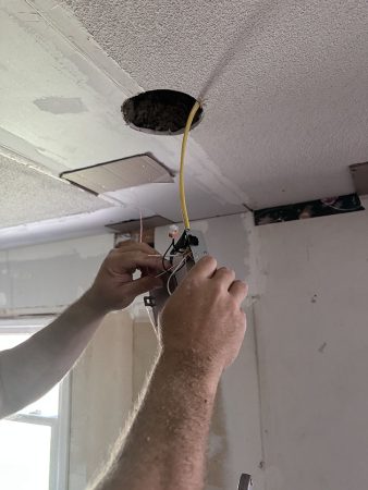A man is repairing a ceiling light fixture in a well-lit room, using tools and standing on a ladder.