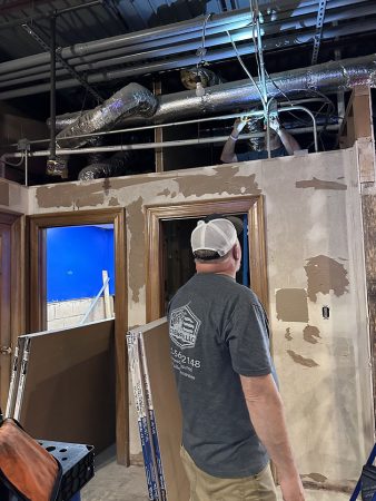 A man stands in a room next to a ladder, looking up as if preparing to climb or work on something above.