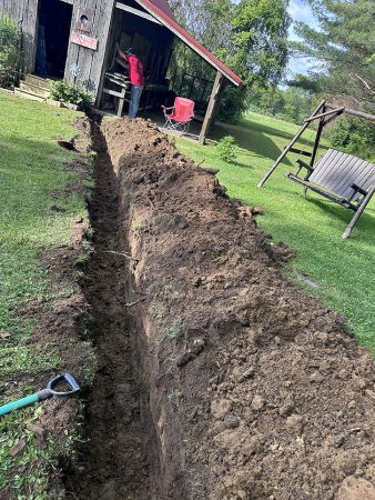 A man is digging a trench in his yard, using a shovel to remove soil.