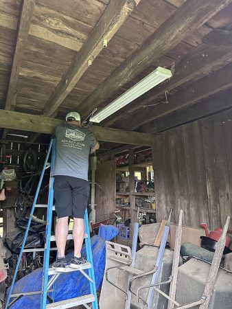 A man is standing on a ladder, working on a project inside a cluttered shed.