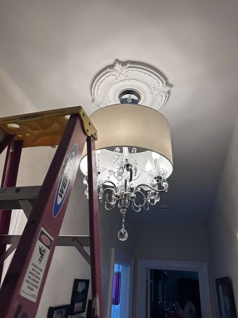 A person on a ladder installs a chandelier in a well-lit room, showcasing the intricate design of the light fixture.