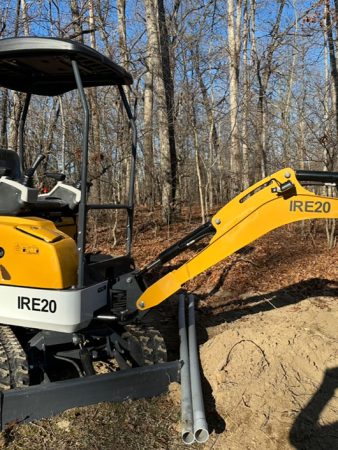 A small excavator parked among trees in a wooded area, surrounded by greenery and natural foliage.