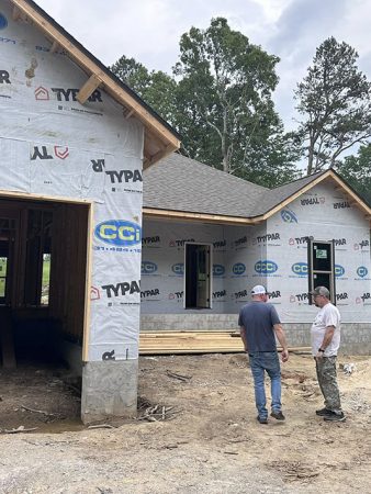 Two men stand in front of a house under construction, discussing the project with building materials visible around them.