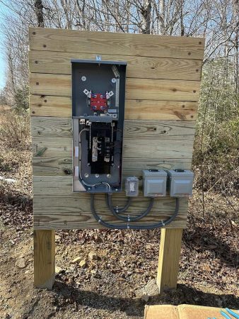 A wooden electrical panel mounted outdoors, featuring a circuit breaker, connectors, and two smaller electrical enclosures.