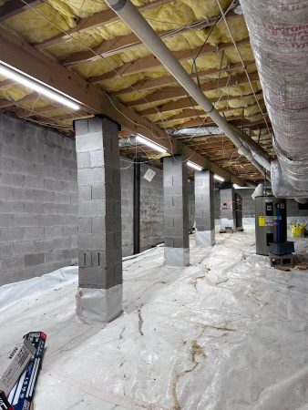 Interior view of a basement under construction, featuring cinderblock columns, plastic flooring, and overhead insulation and lighting.