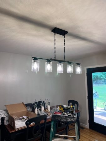 A modern, five-bulb chandelier hangs above a dining table cluttered with tools and decor items, illuminated softy in a home setting.
