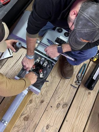 Two men collaboratively working on a wooden piece on a wooden floor, focused on their task.
