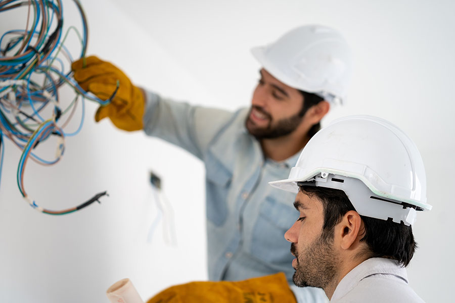 Two construction workers wearing helmets and gloves are handling electrical wires in a bright, minimally furnished room.